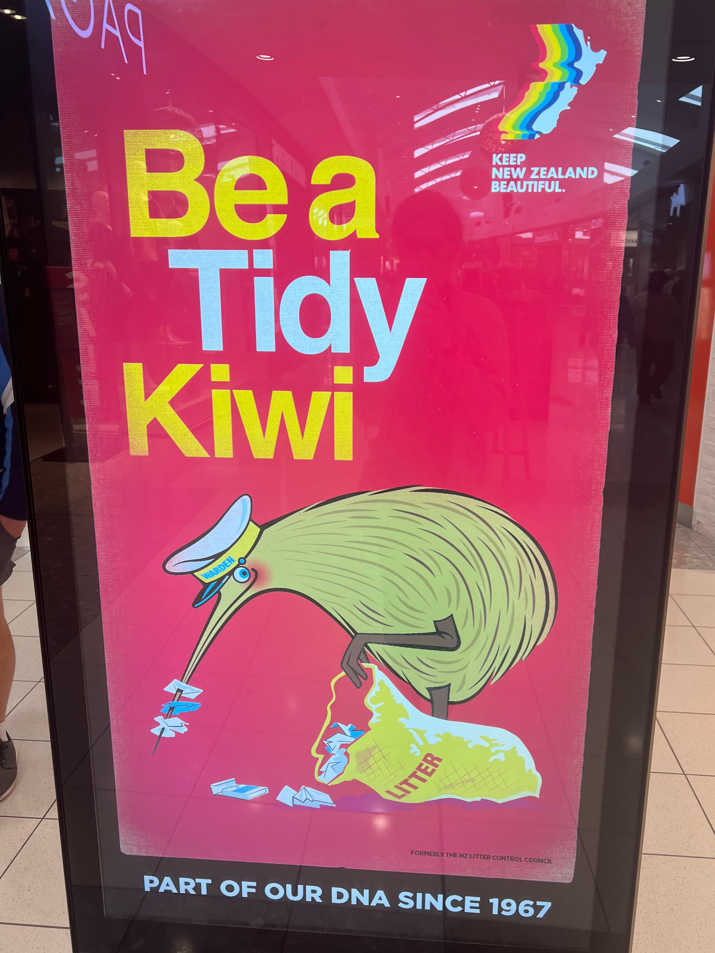 A billboard displaying a kiwi bird dragging a bag of trash with its foot, and using its beak to pick up trash.
